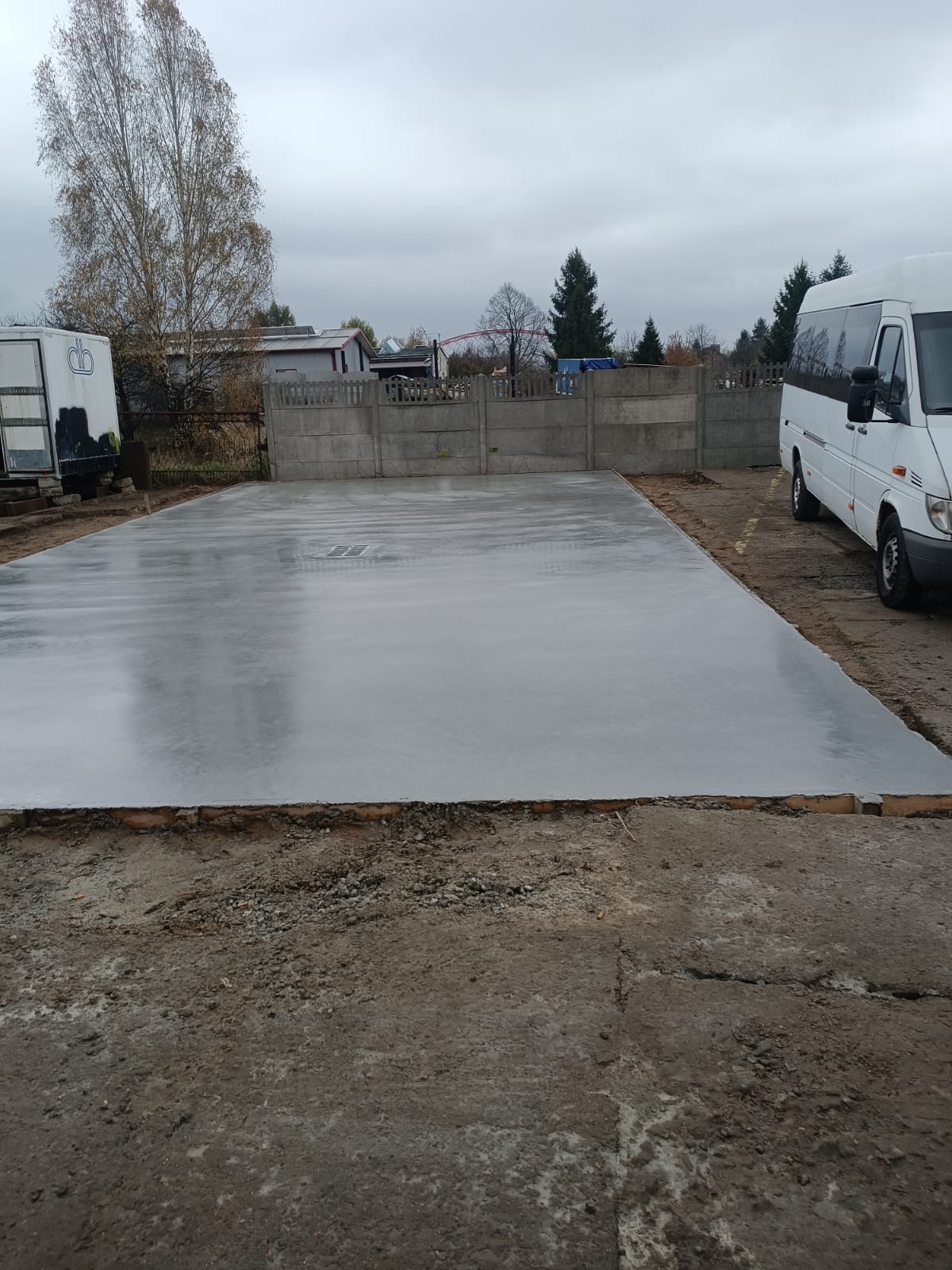 Wylana, gładka, betonowa płyta fundamentowa na działce, z widocznym szalunkiem, obok zaparkowany biały bus, w tle ogrodzenie z prefabrykatów betonowych i drzewa.