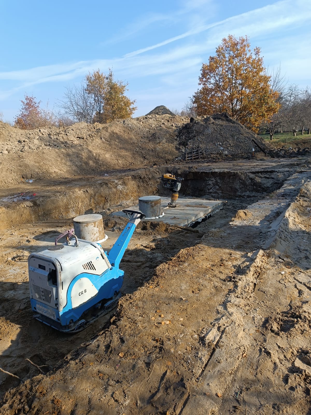 Fundamenty: Wykop z ubijarką CR 8 i wibratorem na betonowej płycie. W tle hałda ziemi i drzewa pod błękitnym niebem.