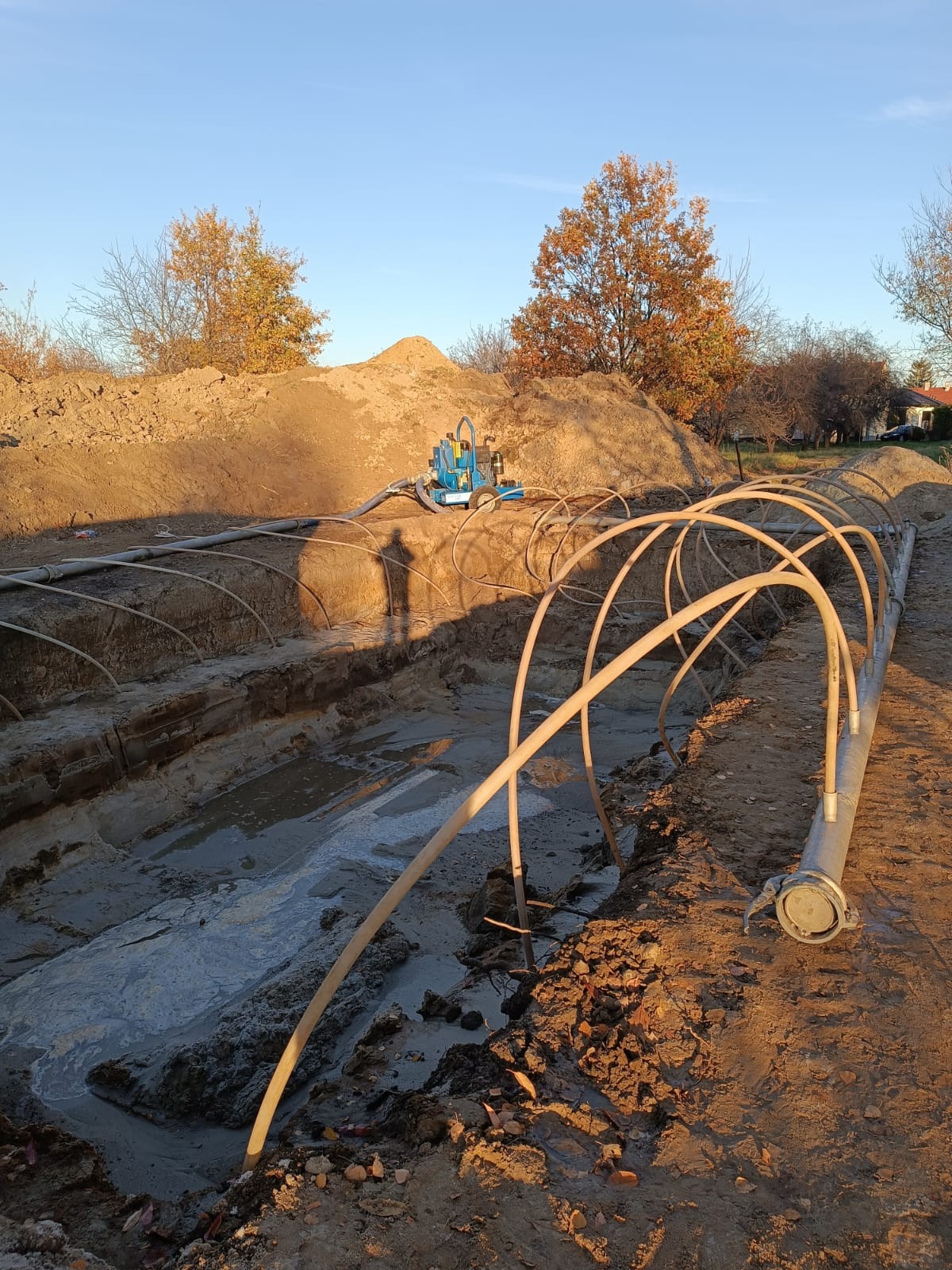 Prace ziemne: wykop z wodą, rury drenażowe ułożone w łuk, maszyna do pompowania wody na tle hałdy ziemi i drzew w jesiennej scenerii.