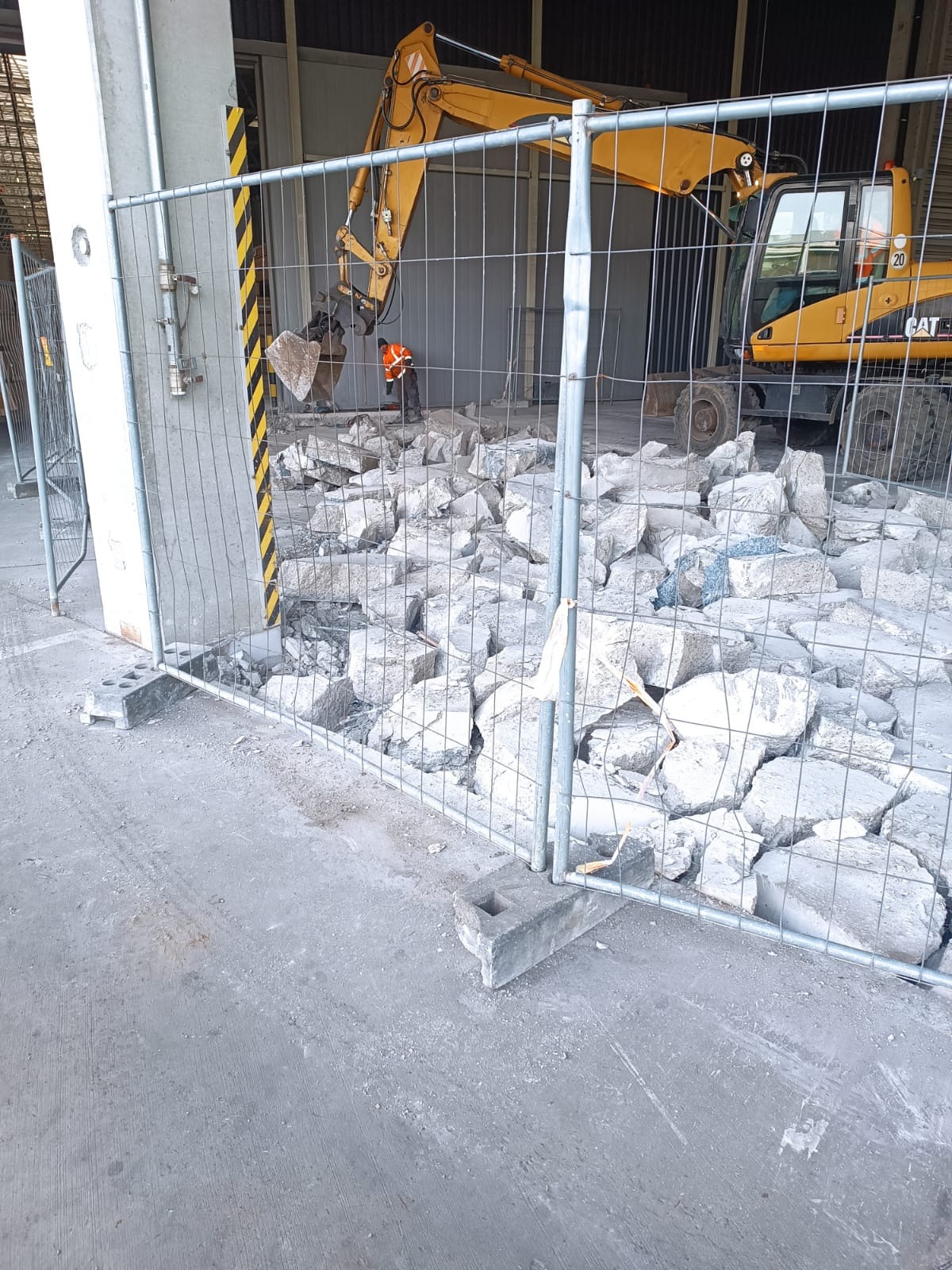 Żółta koparka CAT 20 i robotnik w pomarańczowej kamizelce przy gruzowisku, zabezpieczonym metalowym ogrodzeniem tymczasowym na betonowym podłożu.