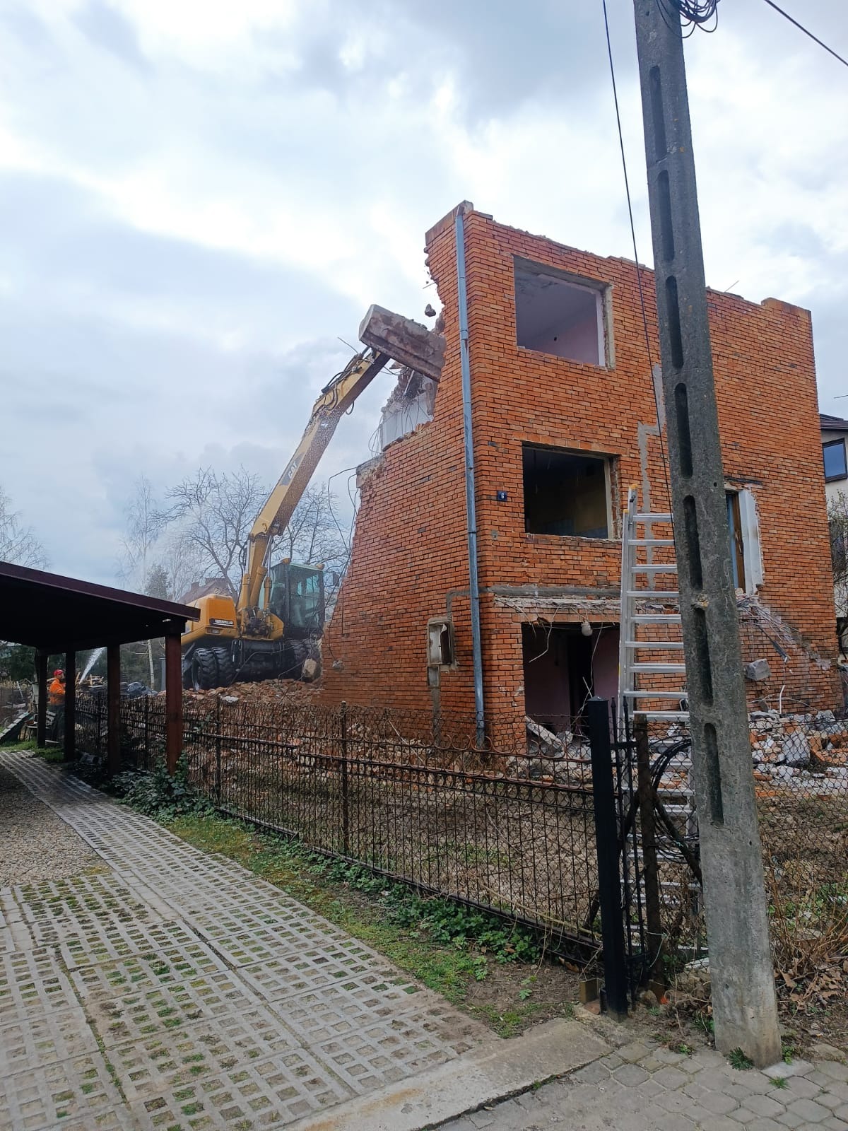 Żółta koparka z hydraulicznym młotem w trakcie rozbiórki ceglanego budynku. Widoczny fragment chodnika z betonowych płyt i słup elektryczny.
