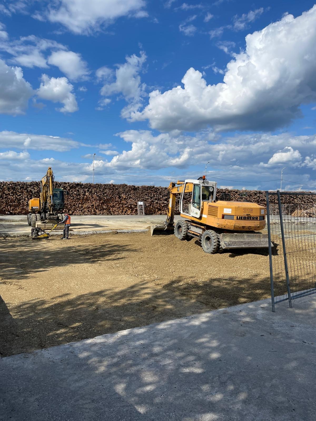 Utwardzanie terenu: Żółta koparka Liebherr wyrównuje ziemię, w tle stos drewna i błękitne niebo z chmurami. Pracownik zagęszcza podłoże ubijarką.