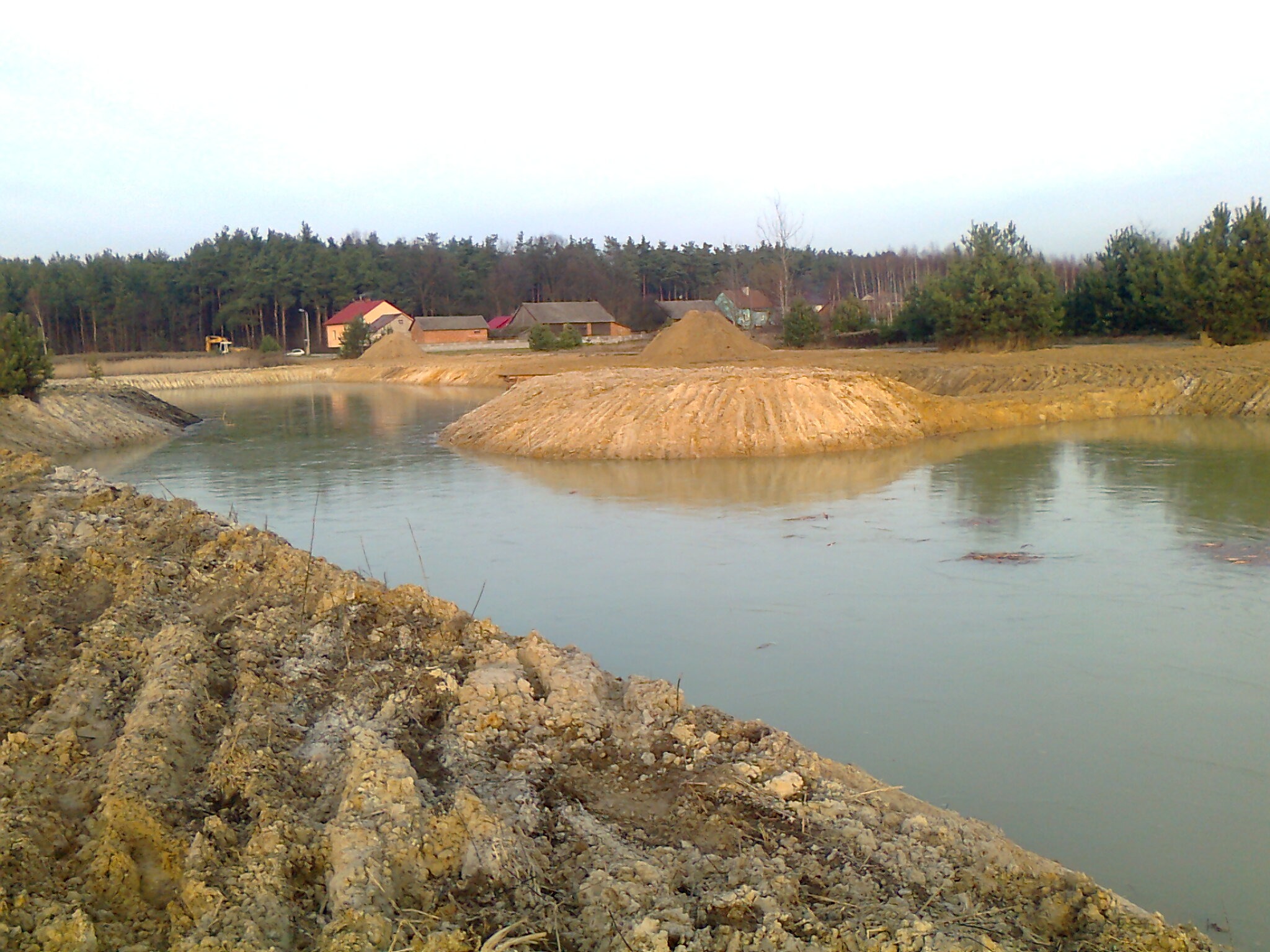 Krajobraz po robotach ziemnych: wykopany staw z wyspą usypaną z ziemi, w tle zabudowania i las. Ślady opon na brzegu.