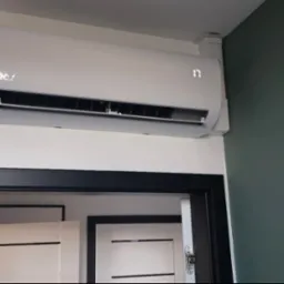 Biały klimatyzator ścienny z wyświetlaczem 17 stopni Celsjusza, zamontowany nad czarną futryną drzwi w nowoczesnym wnętrzu z zieloną ścianą.