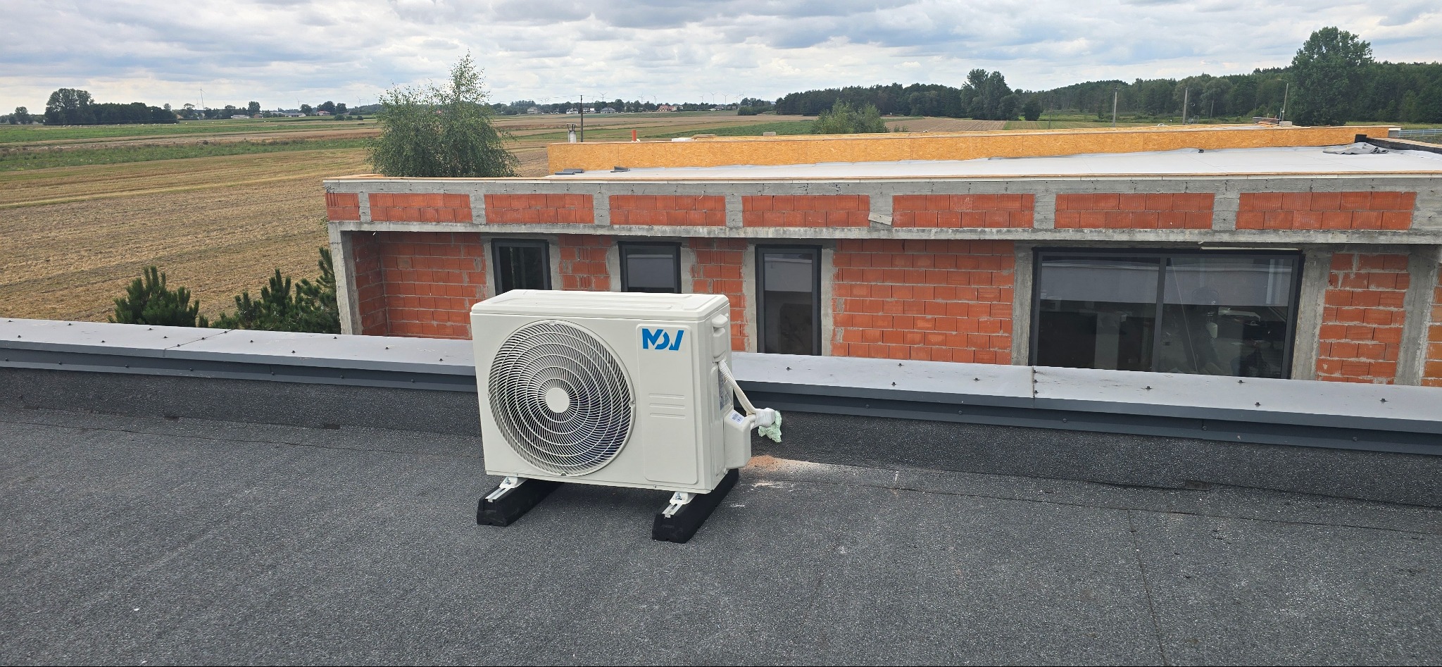 Biała jednostka zewnętrzna klimatyzacji na dachu budynku w trakcie budowy, z widokiem na pola i wiatraki w tle.