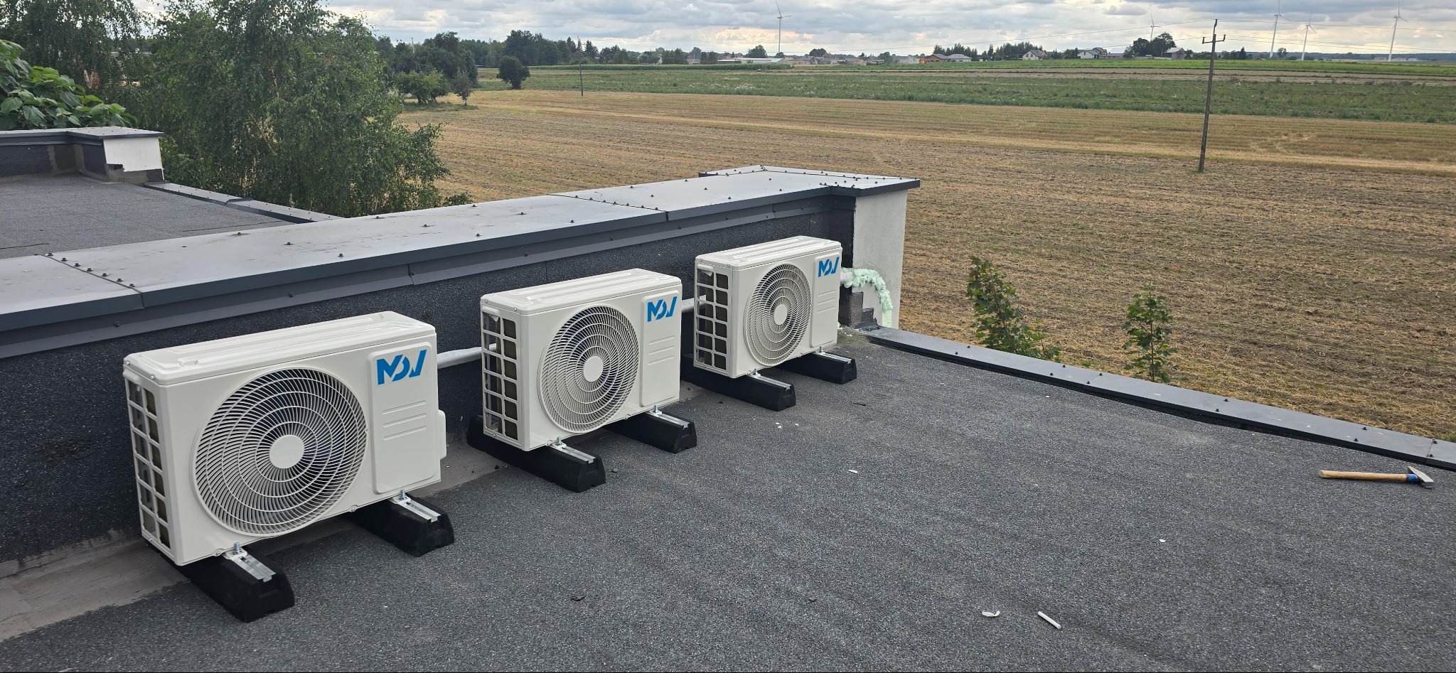 Trzy białe klimatyzatory na czarnym dachu budynku, z widokiem na pola uprawne i turbiny wiatrowe w oddali. Jeden klimatyzator z pianką montażową.