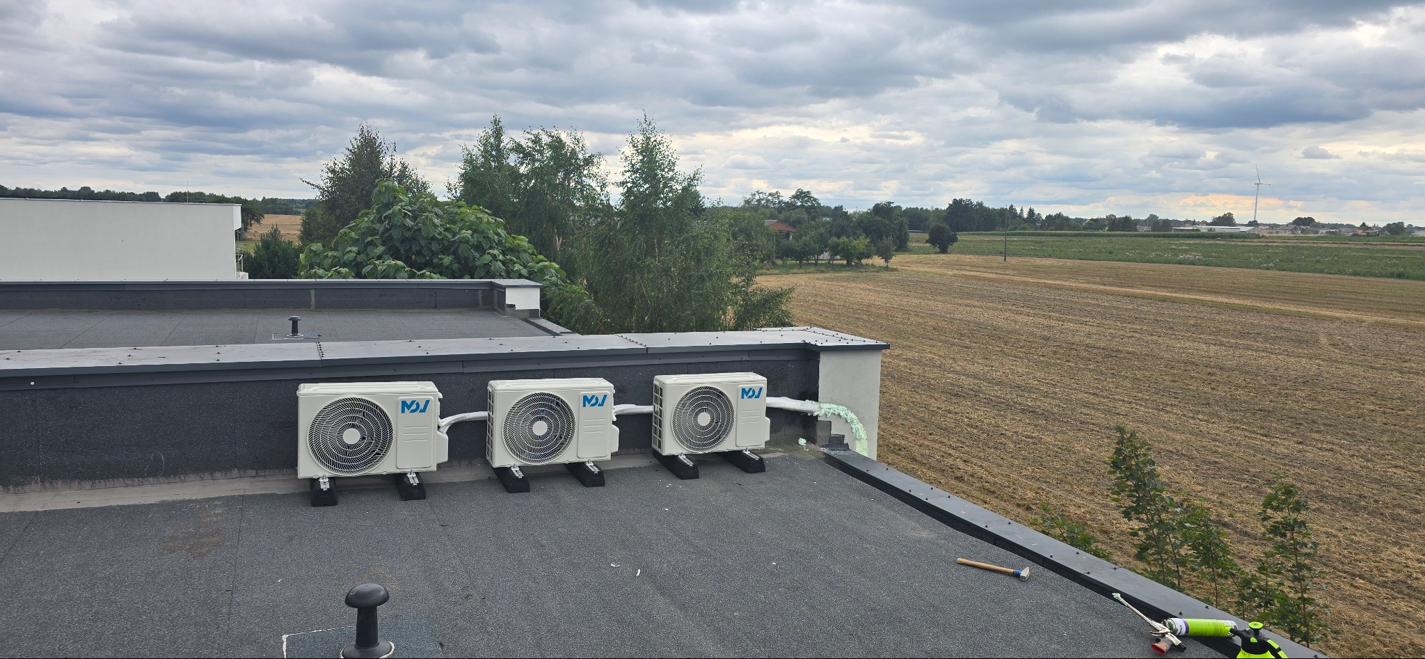 Trzy białe klimatyzatory zamontowane na płaskim dachu budynku z widokiem na pole i wiatrak. Widoczne narzędzia i pianka montażowa.