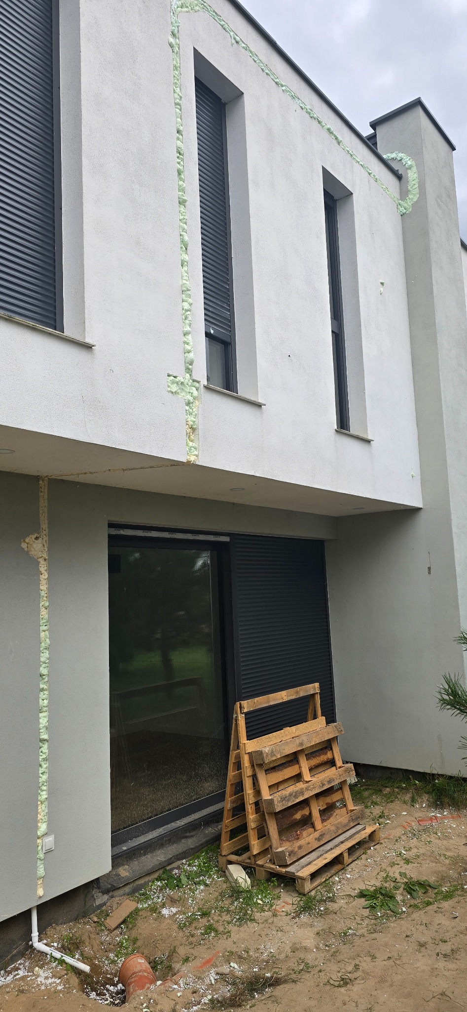 Szczelina dylatacyjna wypełniona pianką montażową na elewacji nowoczesnego domu z szarymi roletami zewnętrznymi. Paleta drewniana przy oknie tarasowym.