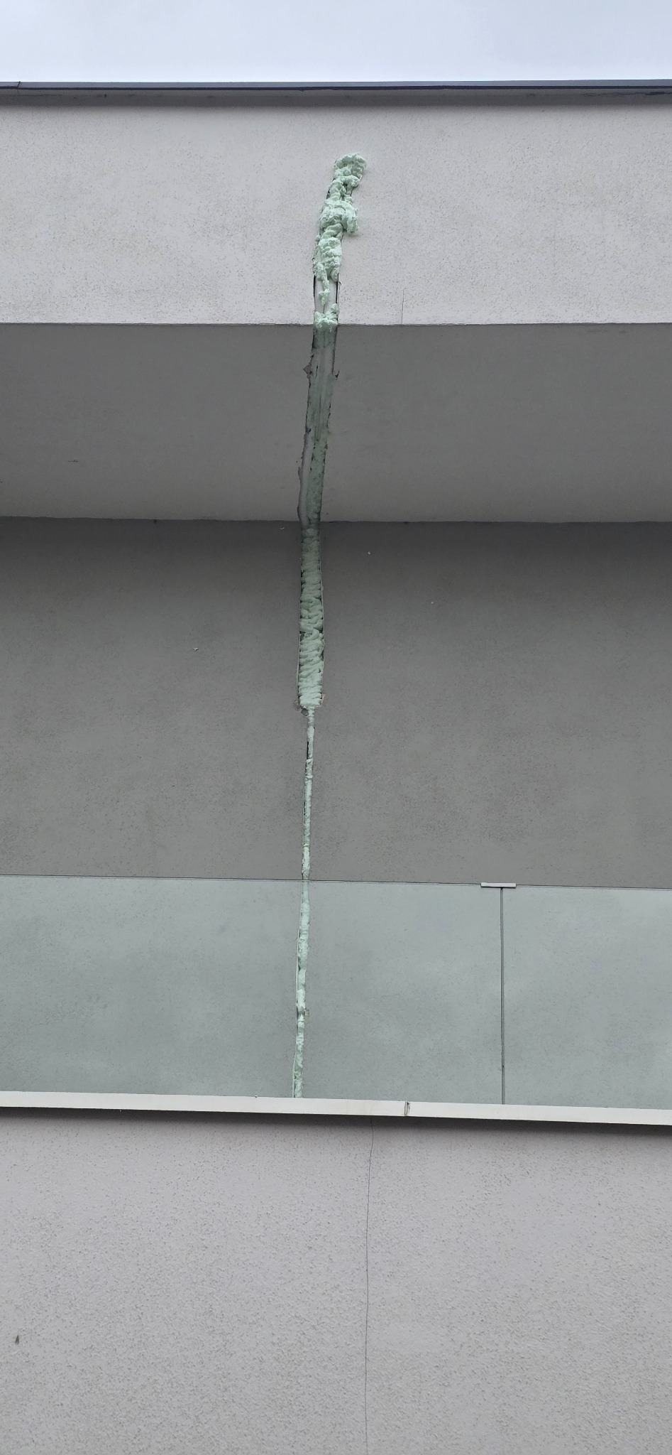 Pionowa szczelina między balkonami budynku wypełniona jasną pianką montażową, widoczna z dołu. Detal naprawy elewacji, szare tło.