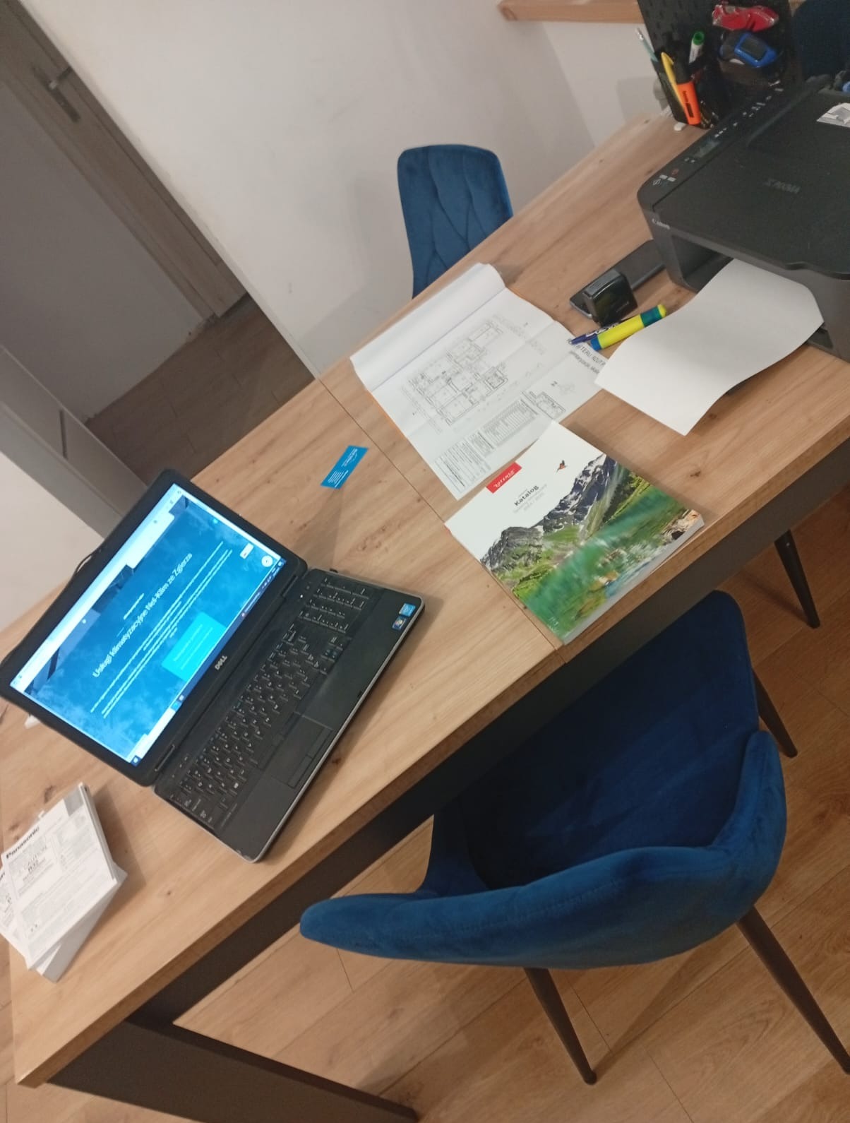 Laptop z otwartą stroną o usługach, plan budynku, katalog i drukarka na drewnianym biurku, sugerujące pracę projektową lub ofertowanie.