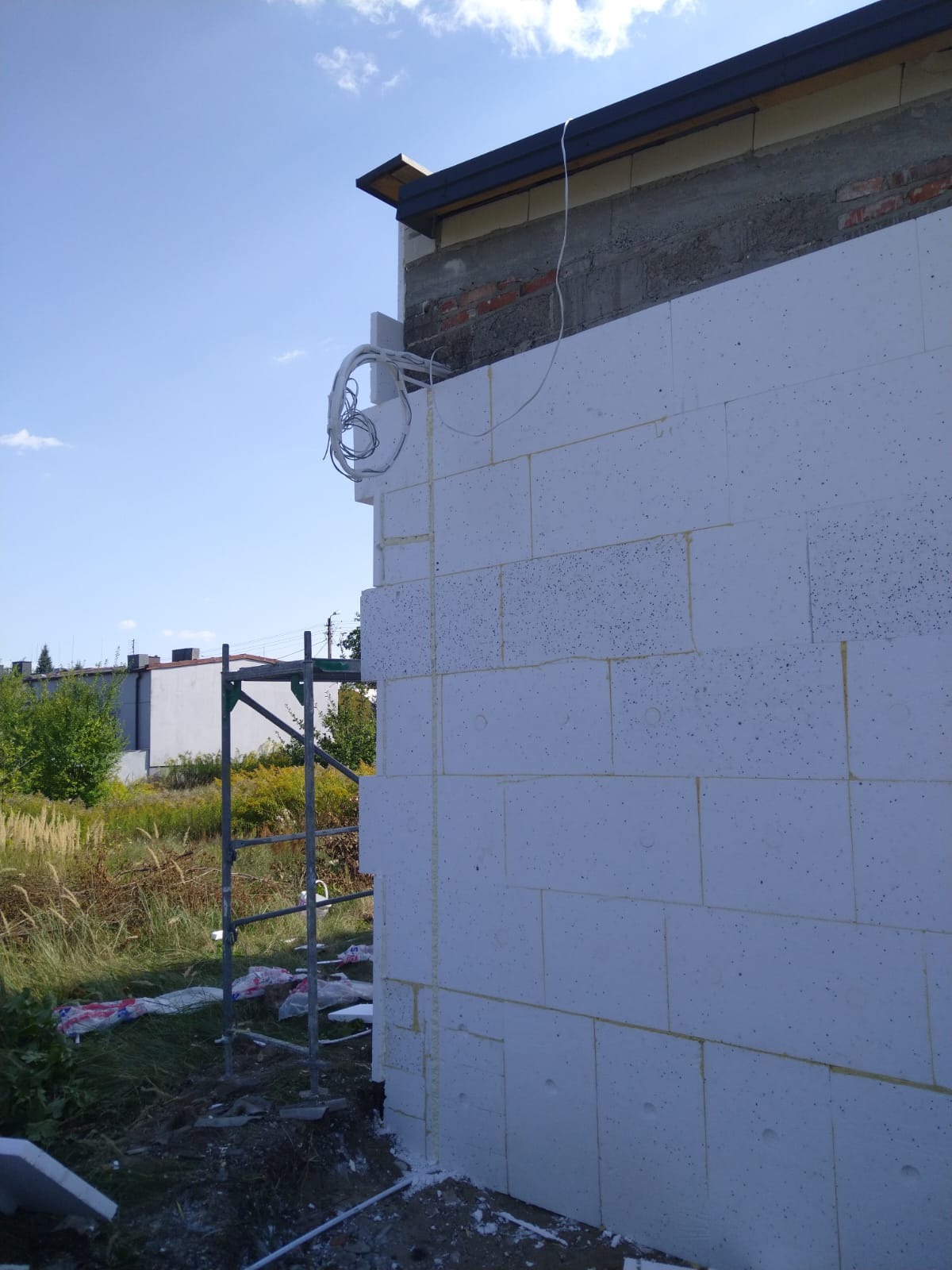 Ściana budynku w trakcie ocieplania styropianem, widoczne przewody elektryczne i rusztowanie. Budowa domu jednorodzinnego w słoneczny dzień.