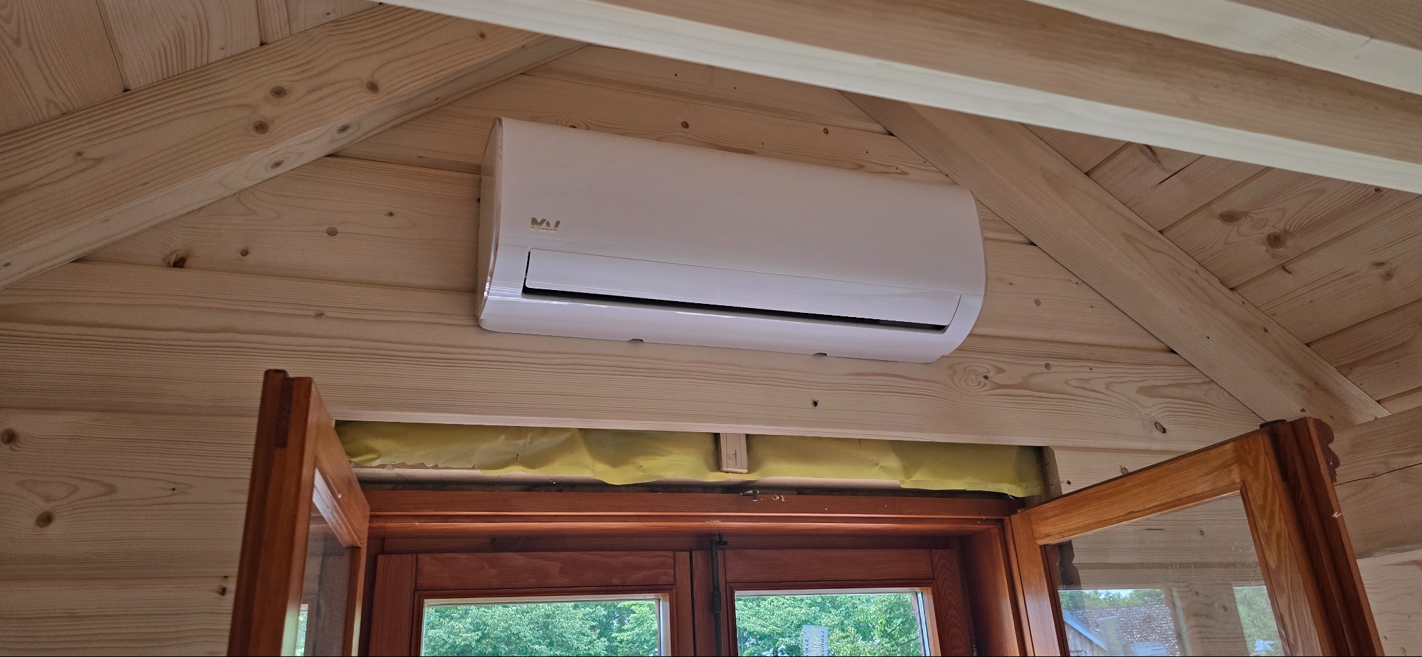 Biały klimatyzator zamontowany na drewnianej ścianie domku, widoczny nad oknem z widokiem na zieleń. Ujęcie od dołu, podkreślające estetykę wnętrza.