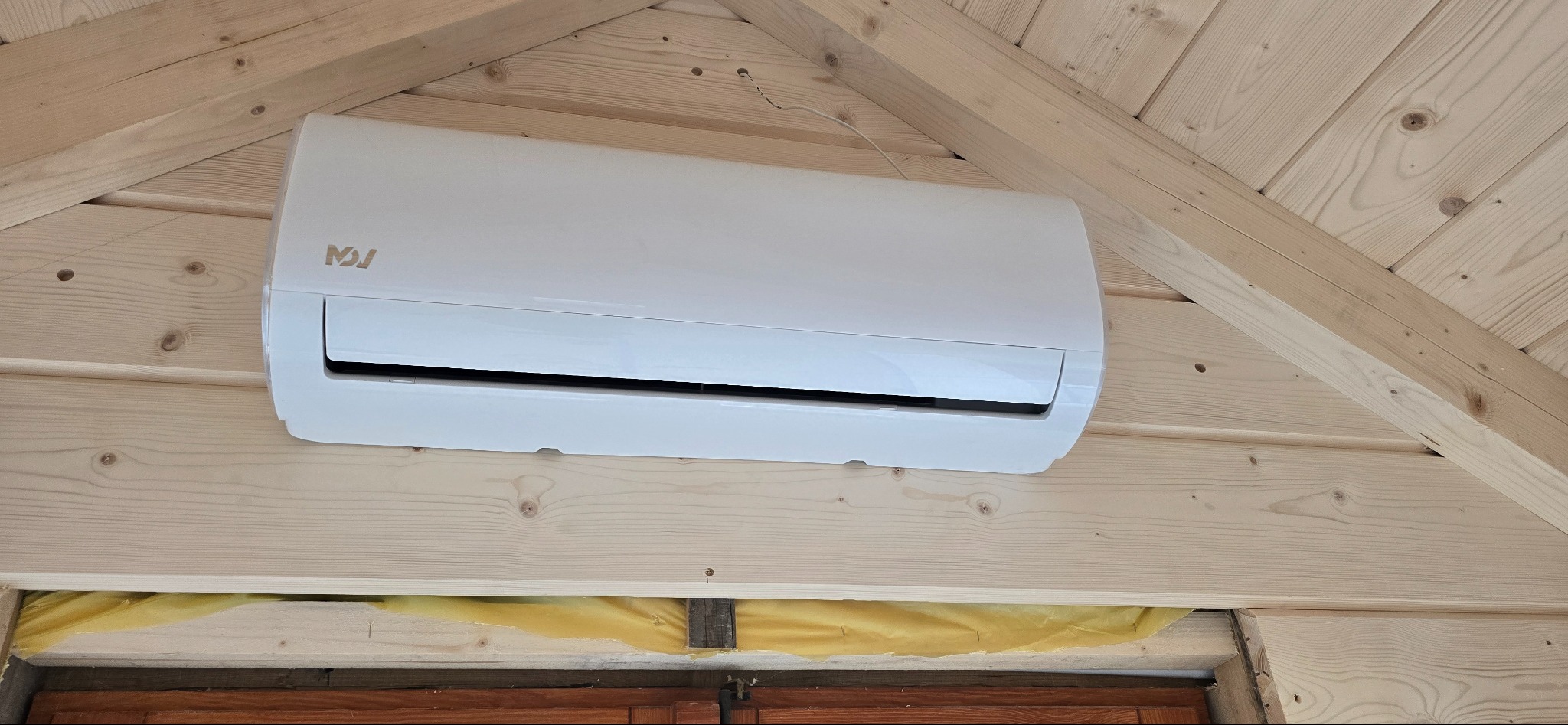 Biała klimatyzacja zamontowana na drewnianej ścianie pod skosem dachu. Widoczne drewniane elementy konstrukcyjne, izolacja. Ujęcie z dołu, z bliska.