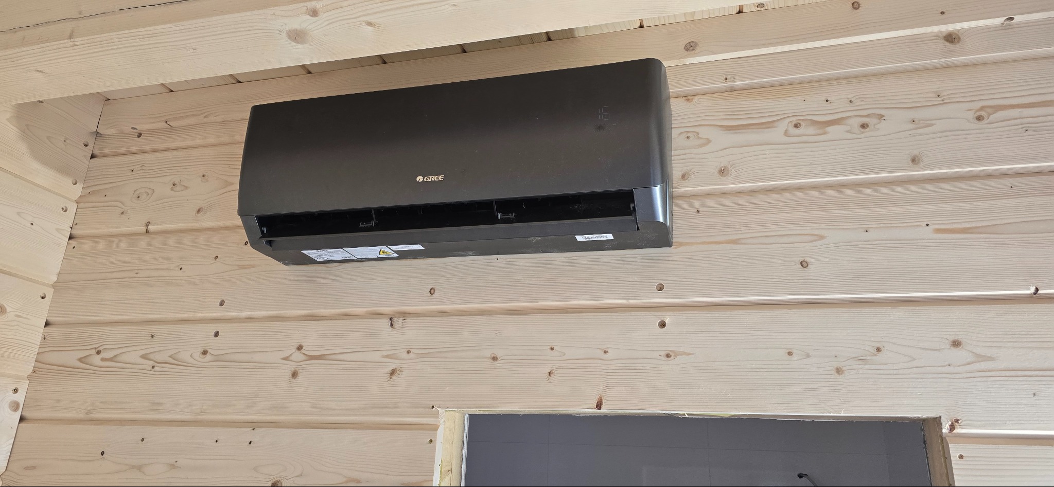 Czarna klimatyzacja Gree zamontowana na jasnej, drewnianej ścianie w domu z bali, widoczny fragment otworu drzwiowego. Nowoczesny design i estetyczny montaż.