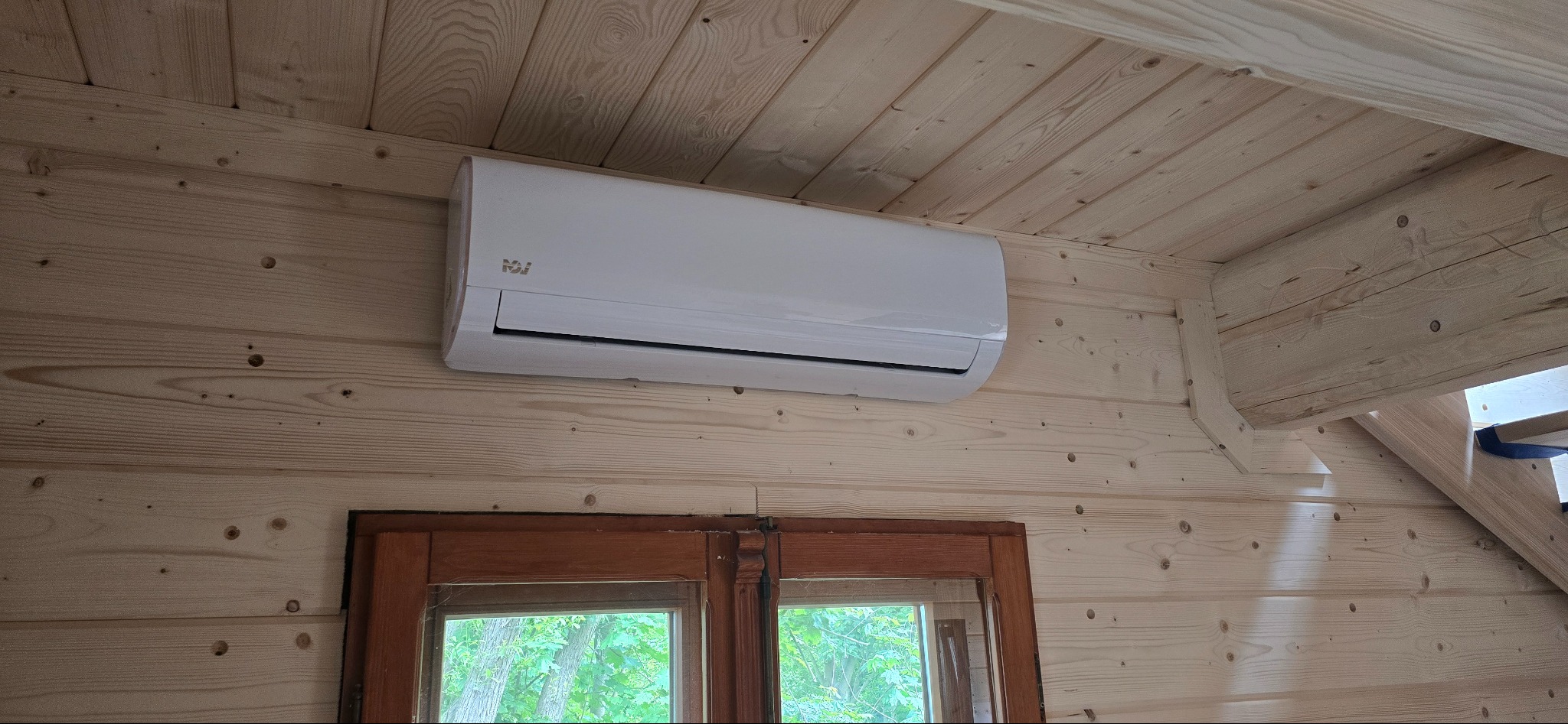 Biały klimatyzator na drewnianej ścianie domku. Widoczne okno z zielenią za szybą. Ujęcie z dołu, podkreślające montaż w drewnianym wnętrzu.