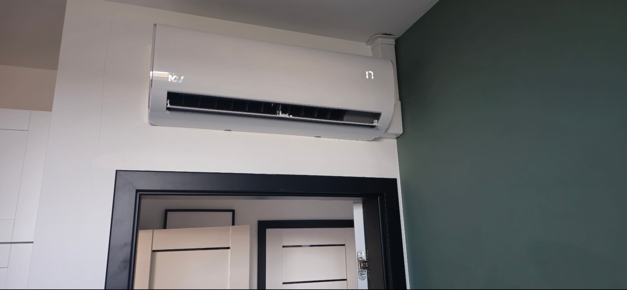 Biały klimatyzator ścienny z wyświetlaczem 17 stopni Celsjusza, zamontowany nad czarną futryną drzwi w nowoczesnym wnętrzu z zieloną ścianą.