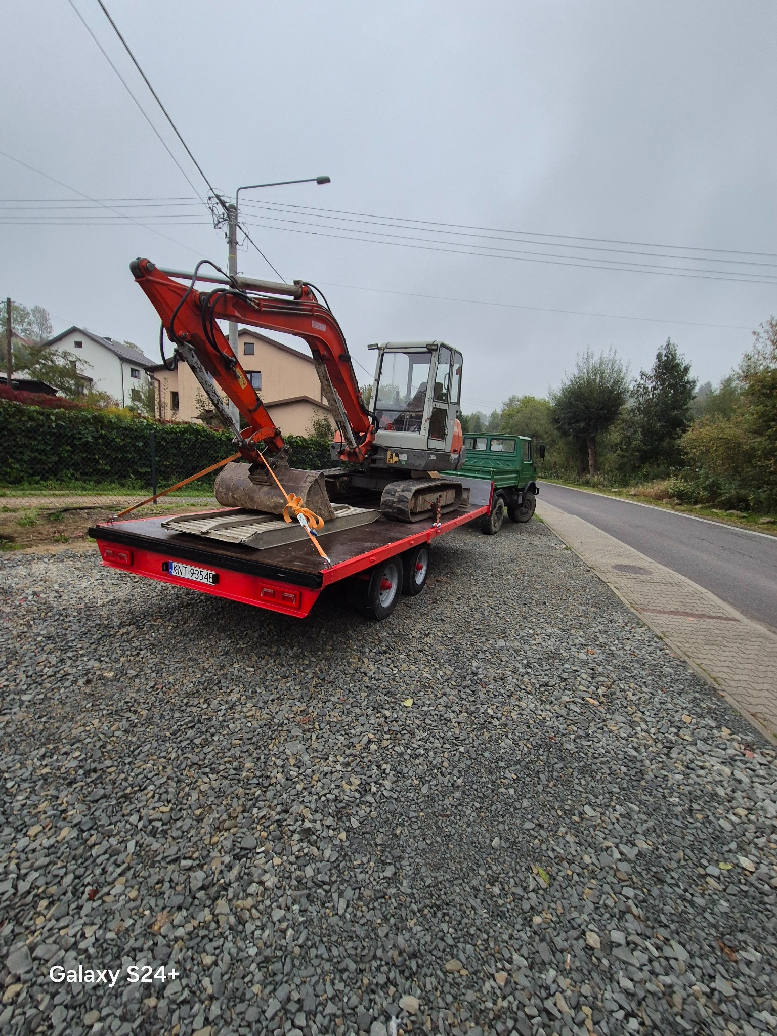 Transport koparki na lawecie w Sieniawie. Czerwona laweta z pomarańczową koparką, zabezpieczona pasami. Z tyłu zielony samochód ciężarowy. Podłoże wysypane kamieniem.
