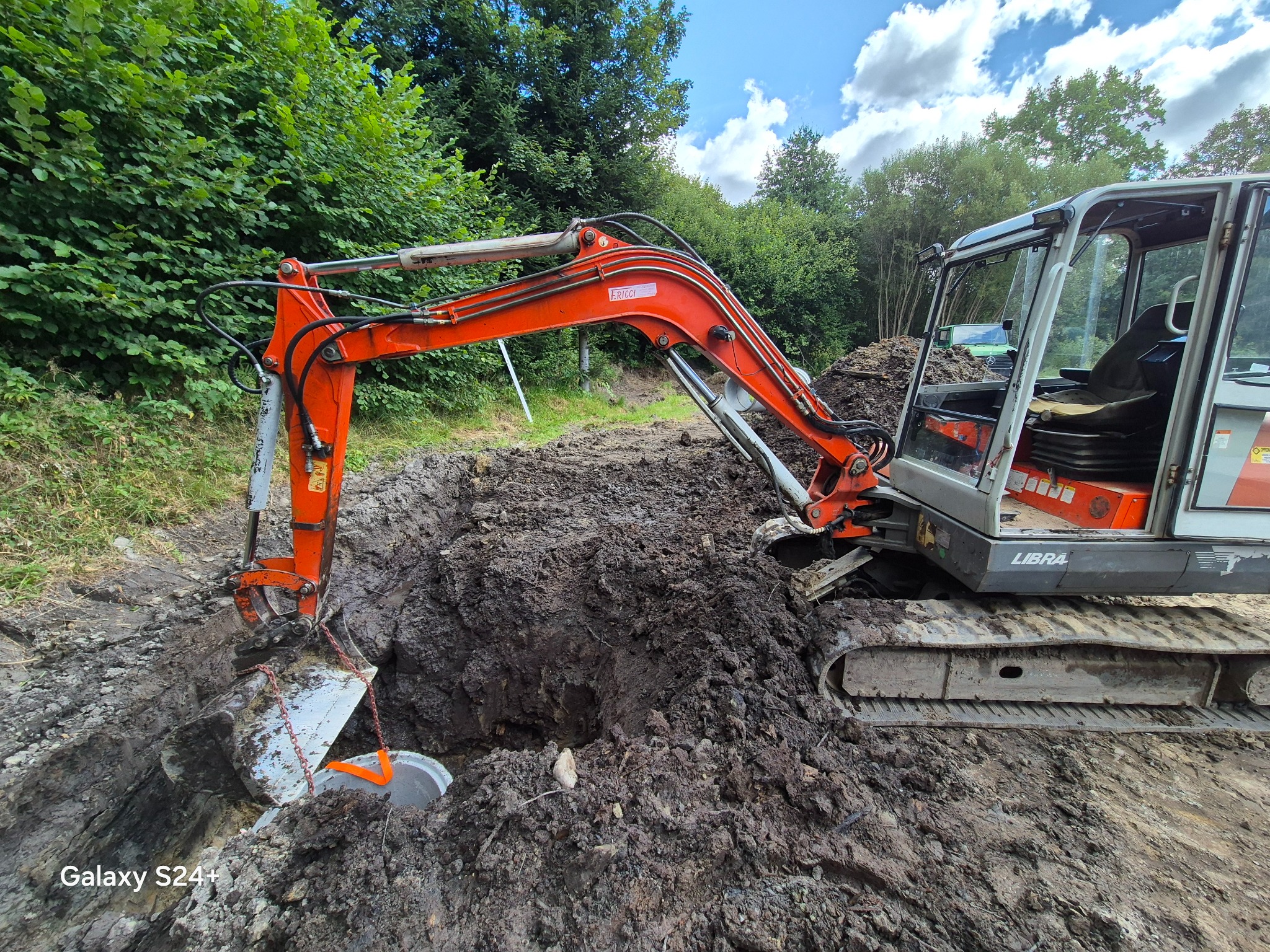 Pomarańczowa koparka RICCI na gąsienicach Libra w trakcie wykopu ziemi, widoczny wykop z szarym betonowym elementem. Tło to drzewa i błękitne niebo.