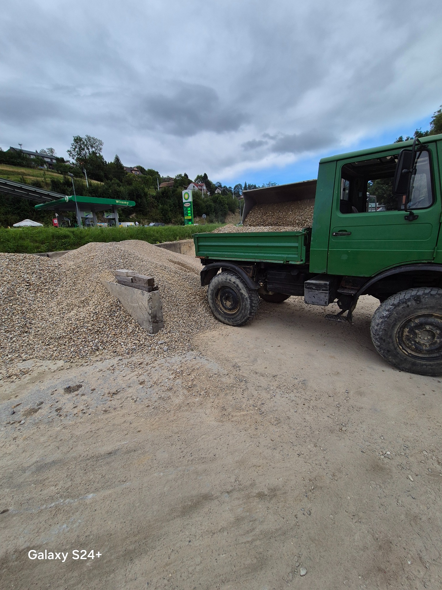 Zielony samochód ciężarowy wyładowuje żwir obok stacji benzynowej, z widocznym stosem żwiru i betonowym obrzeżem. Pochmurne niebo w tle.