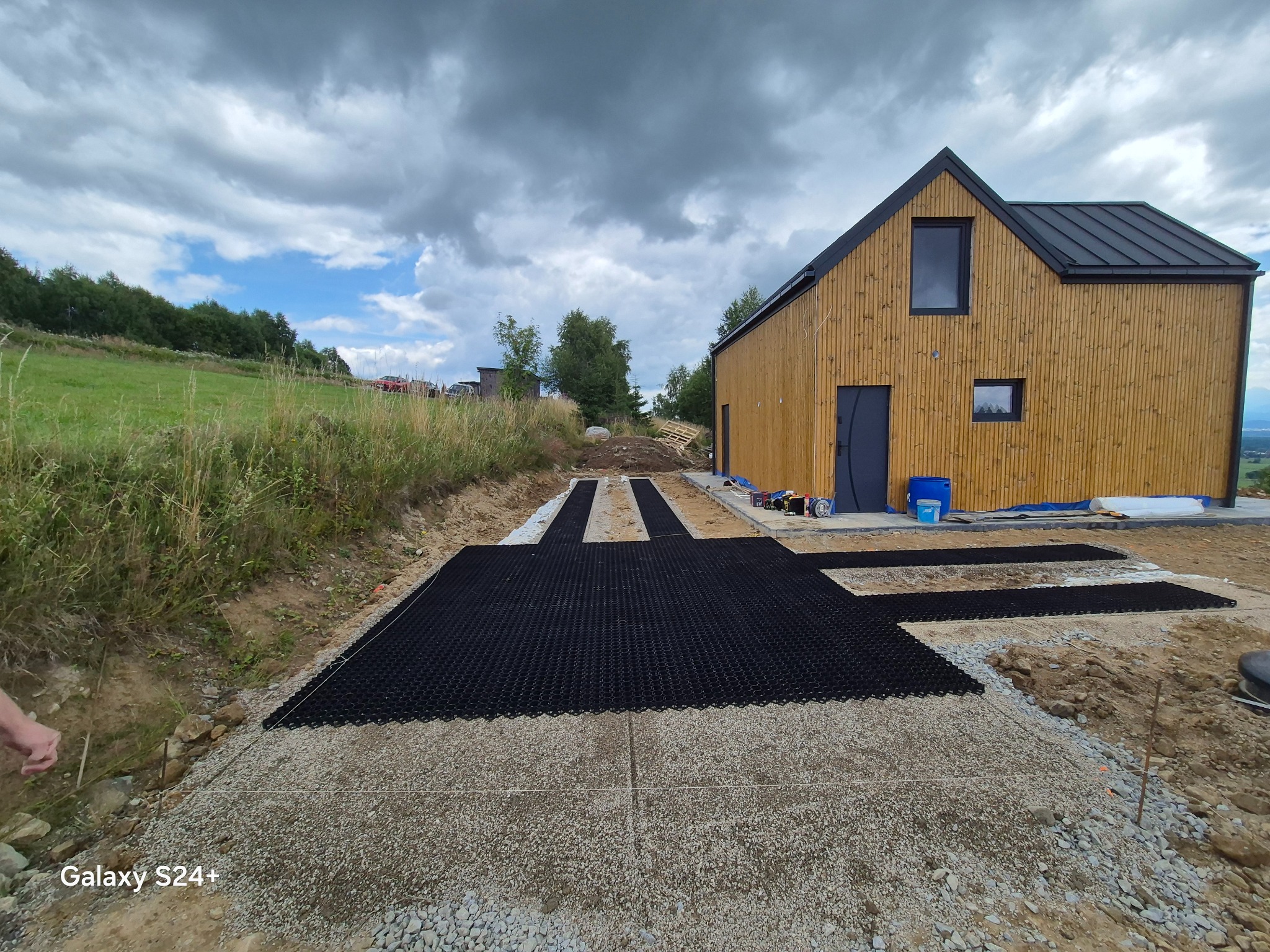 Przygotowanie podłoża z użyciem geokraty na żwirowej podsypce przed domem z drewnianą elewacją, widok na wzgórza i zachmurzone niebo.