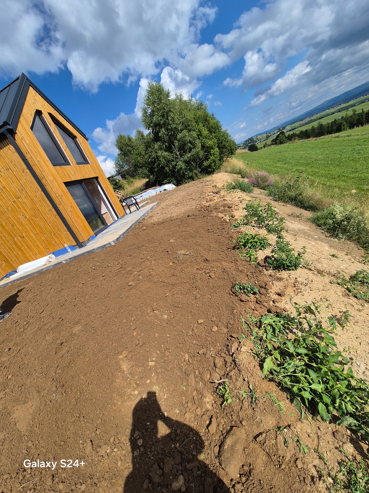 Przygotowany teren pod budowę domu z drewnianą elewacją. W tle zielone wzgórza i błękitne niebo z chmurami. Widoczna granica wyrównanej ziemi i naturalnego zbocza.