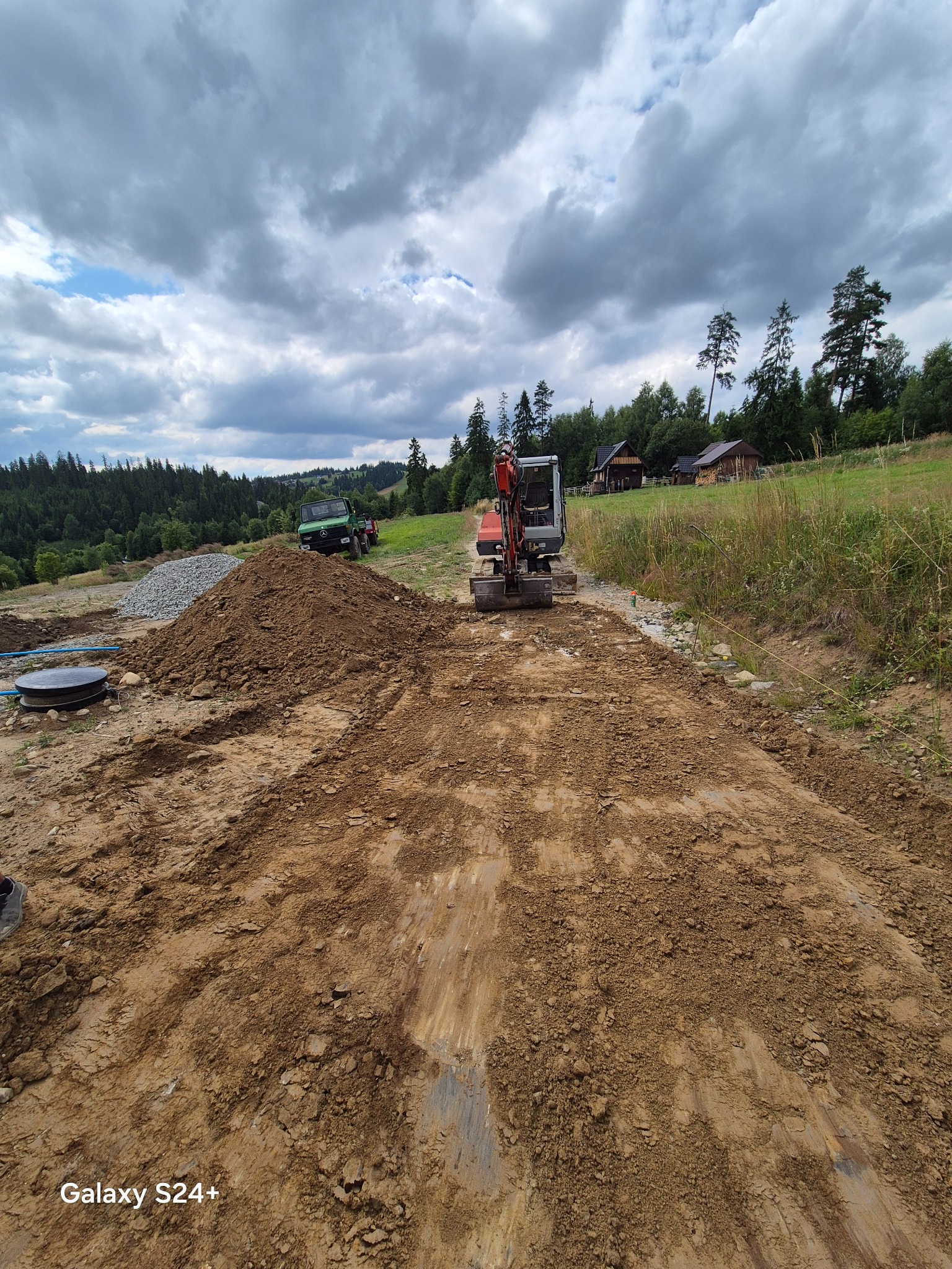 Mini koparka na placu budowy w górskim krajobrazie. Widoczny wykop, hałda ziemi i budynki w tle. Pochmurne niebo nadaje surowy charakter.