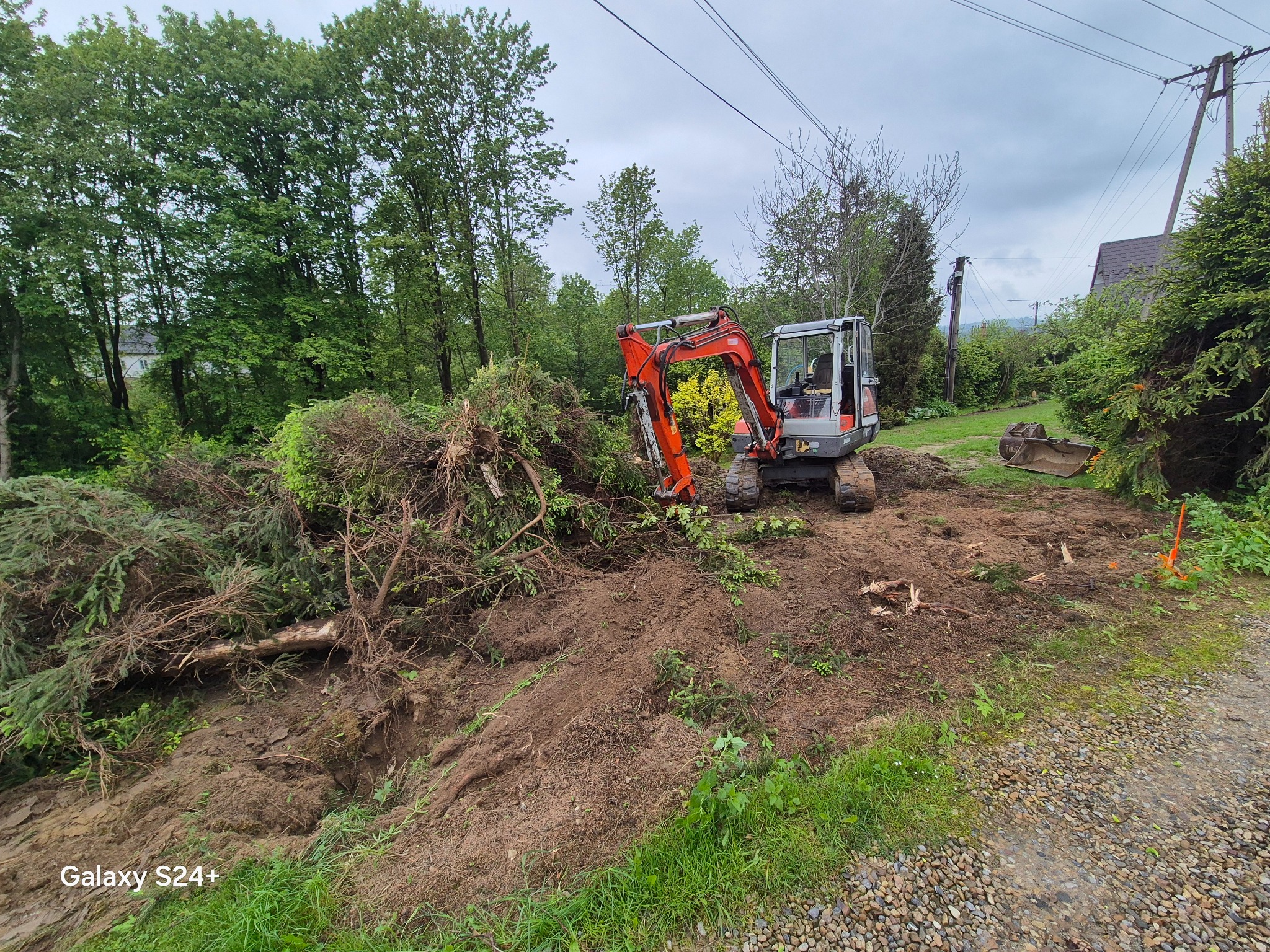 Pomarańczowa mini koparka na placu budowy, hałda ziemi i ścięte gałęzie drzew. Roboty ziemne w toku, widok na zieleń i pochmurne niebo w tle.