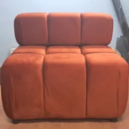 Pomarańczowy, modułowy fotel tapicerowany w stylu retro, na drewnianym blacie warsztatowym. W tle widoczna szafka z szufladami i piec.