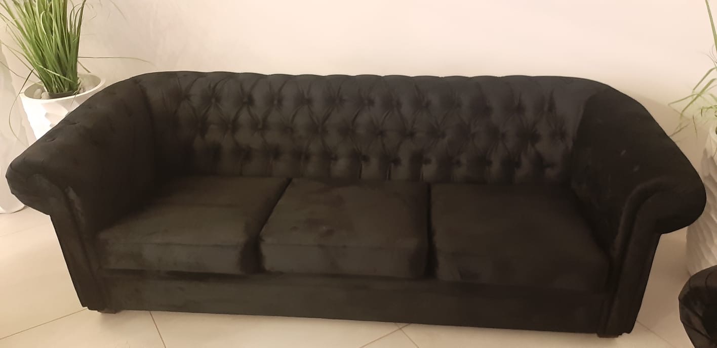 Czarna, welurowa sofa Chesterfield z pikowanym oparciem, ustawiona na jasnej podłodze, z roślinami doniczkowymi po bokach. Elegancki mebel w neutralnym wnętrzu.