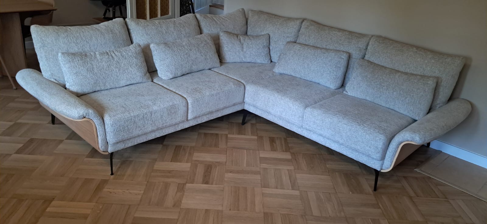 Jasnoszara sofa narożna z poduszkami, drewnianymi elementami i metalowymi nóżkami na parkiecie. Nowoczesny design, stonowana kolorystyka i minimalistyczny styl.