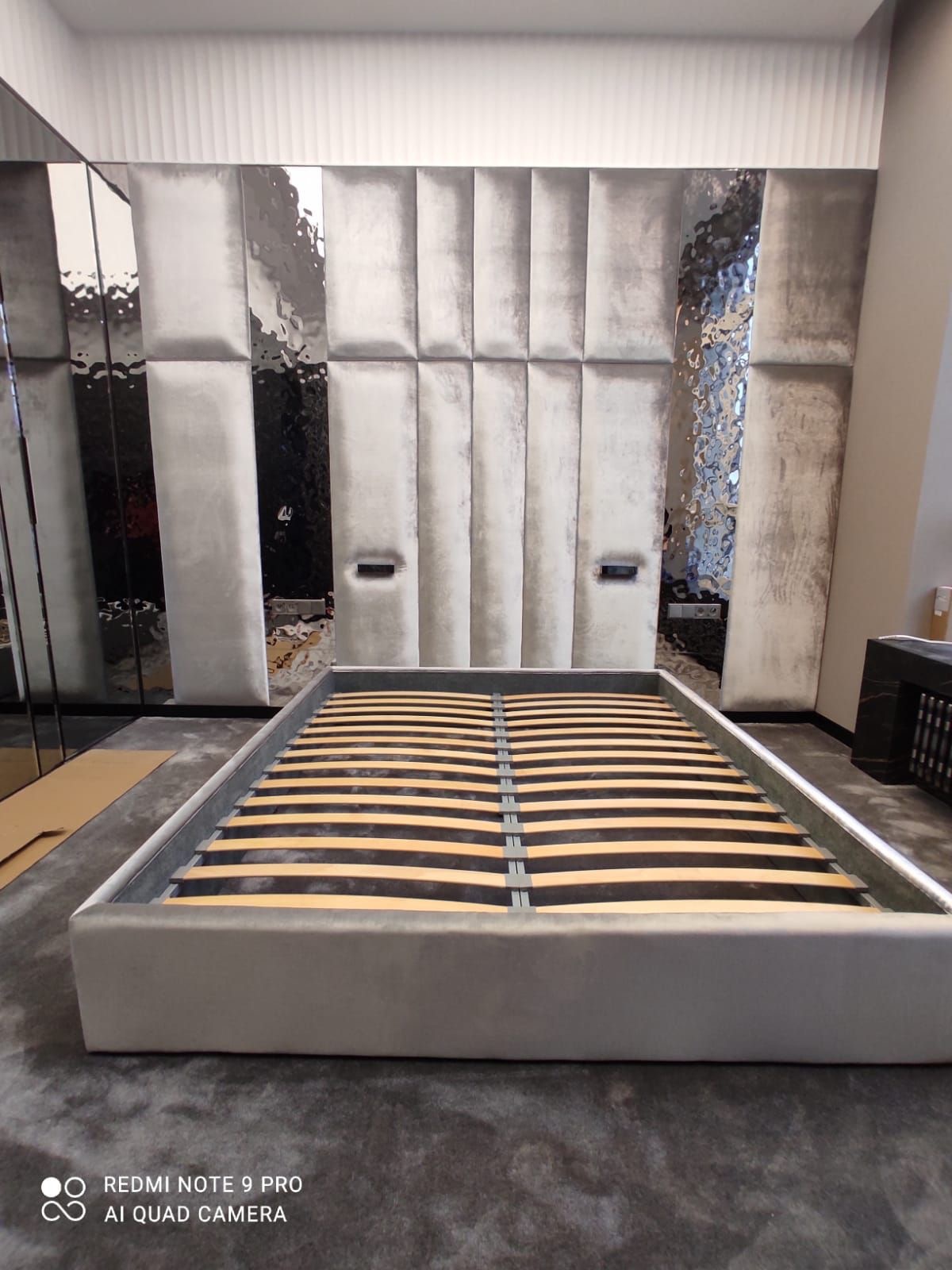 Szare, tapicerowane łóżko z wezgłowiem z panelami i lustrami. Widoczna drewniana rama z listwami. Minimalistyczny design, nowoczesny styl.