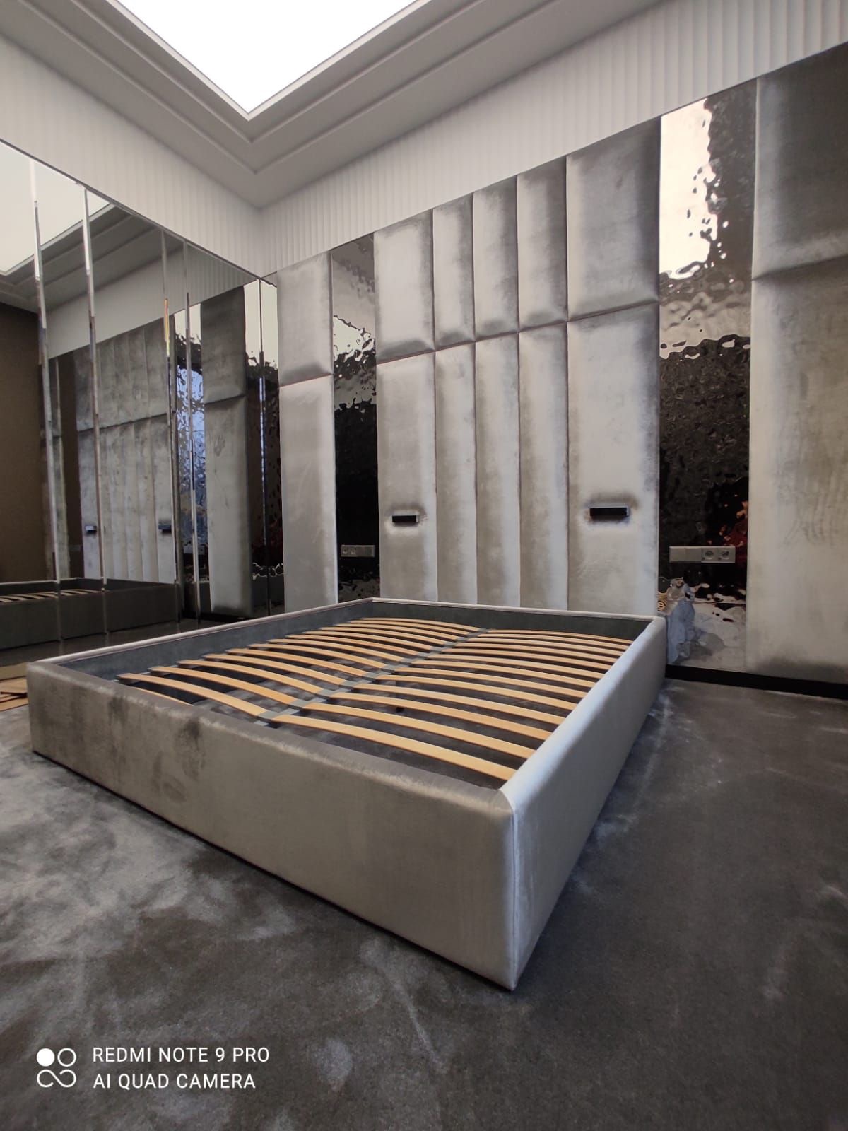 Szare, tapicerowane łóżko z drewnianym stelażem na tle ściany z panelami i lustrami. Nowoczesny design sypialni z elementami odbijającymi światło.