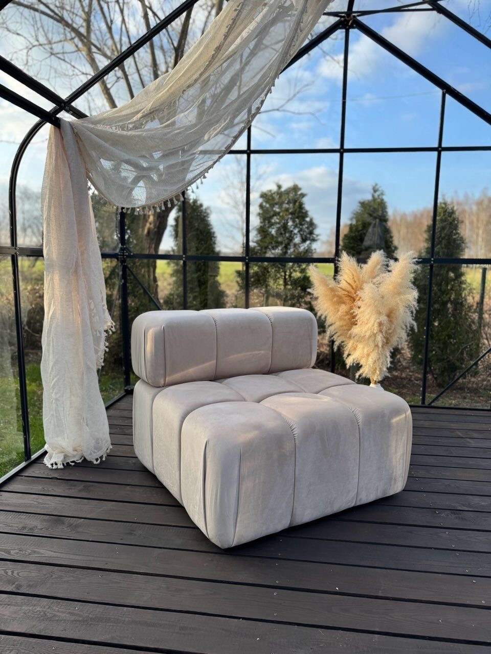 Beżowy, modułowy fotel tapicerowany w szklanej oranżerii z drewnianą podłogą. W tle widok na zieleń i błękitne niebo.