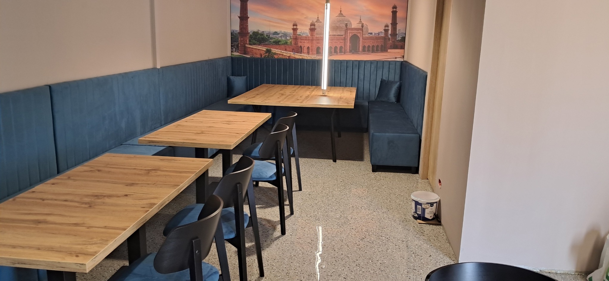 Narożna, błękitna sofa tapicerowana z drewnianymi stolikami i krzesłami w lokalu gastronomicznym, na ścianie fototapeta z motywem architektonicznym.