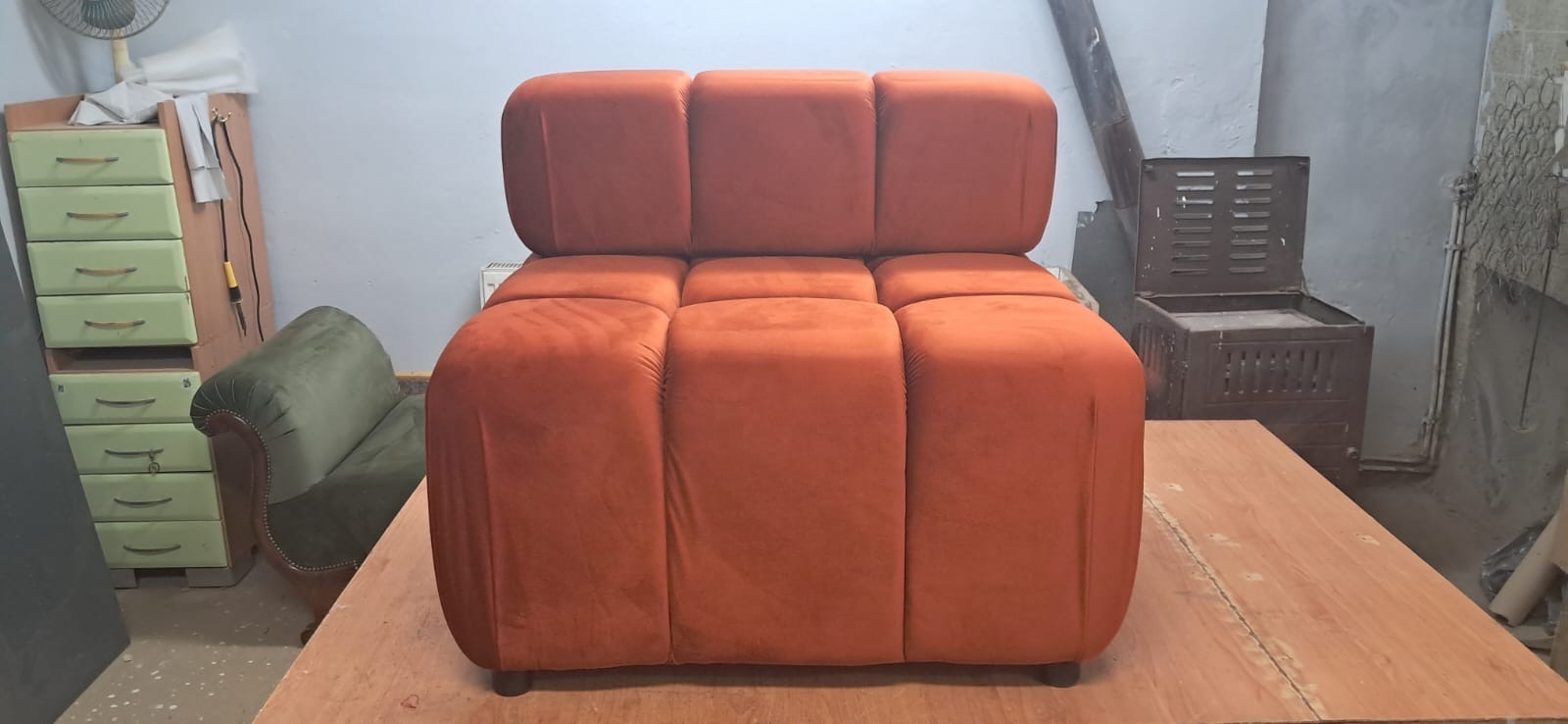 Pomarańczowy, modułowy fotel tapicerowany w stylu retro, na drewnianym blacie warsztatowym. W tle widoczna szafka z szufladami i piec.