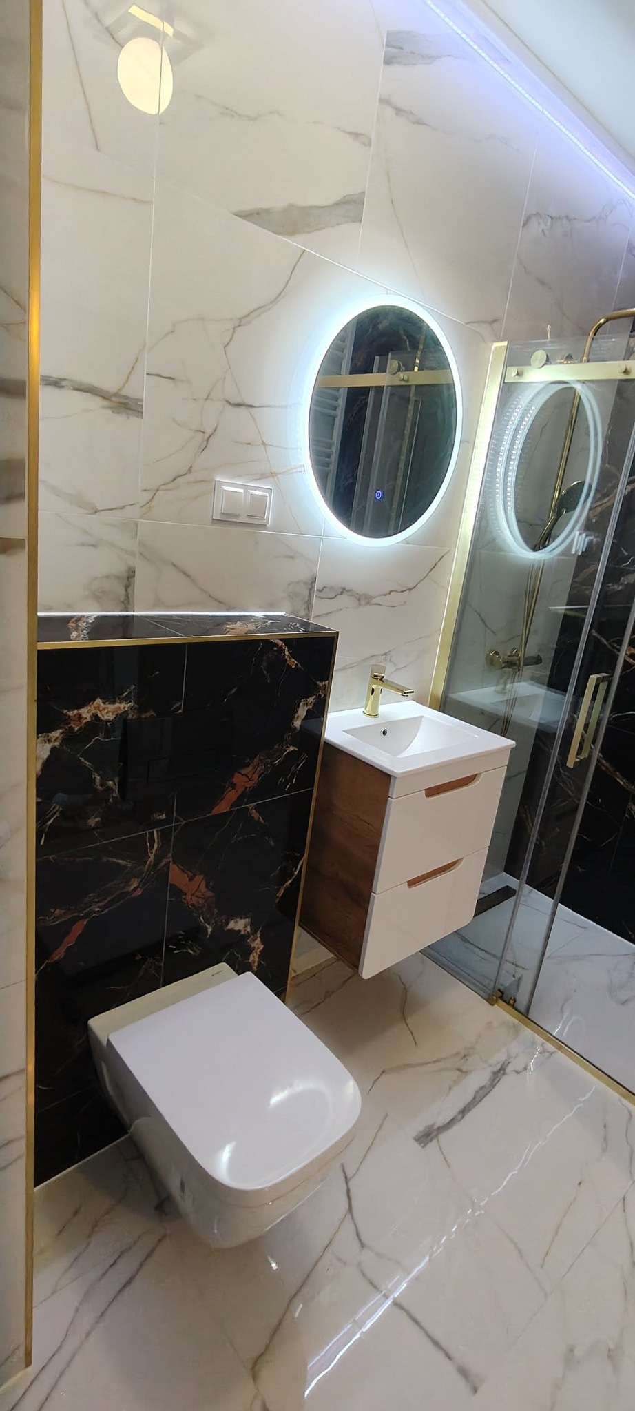 Elegancka łazienka z marmurowymi płytkami, okrągłymi lustrami z podświetleniem LED i kabiną prysznicową ze złotymi detalami.