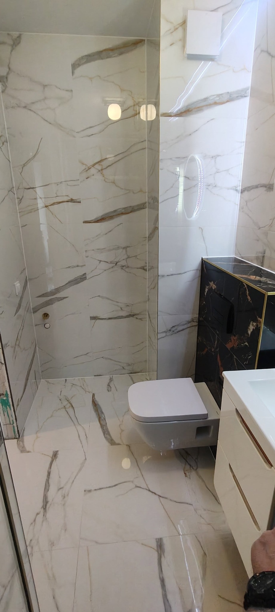 Elegancka łazienka z marmurowymi płytkami, podwieszaną toaletą i szafką umywalkową w kolorze białym, oświetlona ledowym lustrem.