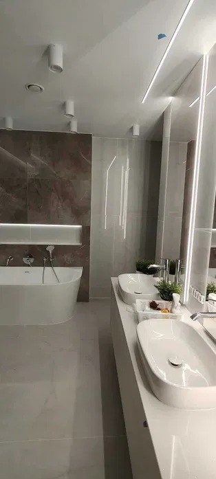 Nowoczesna łazienka z dwoma umywalkami na białym blacie, marmurową ścianą i podświetlaną wnęką nad wanną. Minimalistyczne oświetlenie sufitowe.