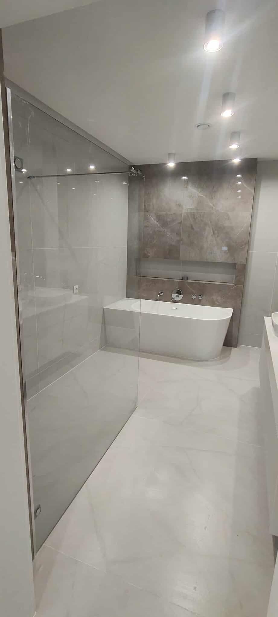 Elegancka łazienka z kabiną prysznicową ze szkła i wolnostojącą wanną na tle ściany z płytek imitujących marmur, oświetlona punktowo.