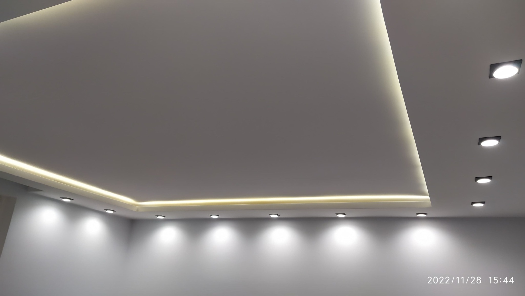 Nowoczesny sufit z oświetleniem LED wzdłuż krawędzi i punktowymi lampami wpuszczanymi, tworzący geometryczny wzór na gładkiej, jasnej powierzchni.