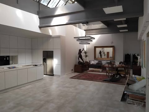 Przestronne wnętrze z nowoczesną, białą zabudową kuchenną, szarymi płytkami podłogowymi i elementami dekoracyjnymi. Widok na część jadalną i salon. Duże okno dachowe.