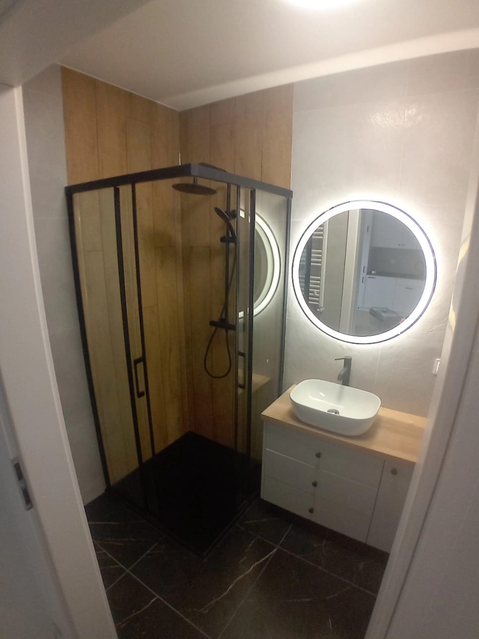 Nowoczesna łazienka z czarną kabiną prysznicową typu walk-in, umywalką nablatową i okrągłym lustrem z podświetleniem LED.