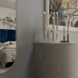 PORADKO DESIGN - Sypialnia z niebieskim zagłówkiem łóżka, widziana w odbiciu lustra. Na komodzie świeczniki i obraz. Stonowana kolorystyka, nowoczesny design.