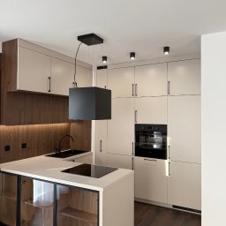 Budservice - Nowoczesna kuchnia z wyspą, fronty w odcieniach beżu i drewna, czarne akcenty: bateria, okap, piekarnik. Oświetlenie punktowe podkreśla minimalistyczny design.