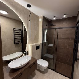 Budservice - Nowoczesna łazienka z kabiną prysznicową typu walk-in, umywalką nablatową na drewnianej szafce i okrągłym lustrem z podświetleniem LED. Beżowo-brązowa kolorystyka.