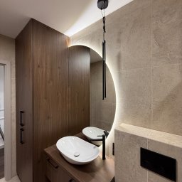 Budservice - Nowoczesna łazienka z umywalką nablatową, okrągłym lustrem z podświetleniem LED i szafką z ciemnego drewna. Beżowe płytki na ścianach i podłodze.