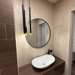 Budservice - Nowoczesna łazienka z owalną umywalką, okrągłym lustrem i wiszącymi lampami. Beżowe płytki na ścianach i drewnopodobna szafka pod umywalką tworzą spójny design.
