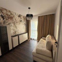 Budservice - Elegancki salon z sofą, komodą i fototapetą w kwiaty. Ciemna podłoga i nowoczesny żyrandol. Stonowana kolorystyka wnętrza.