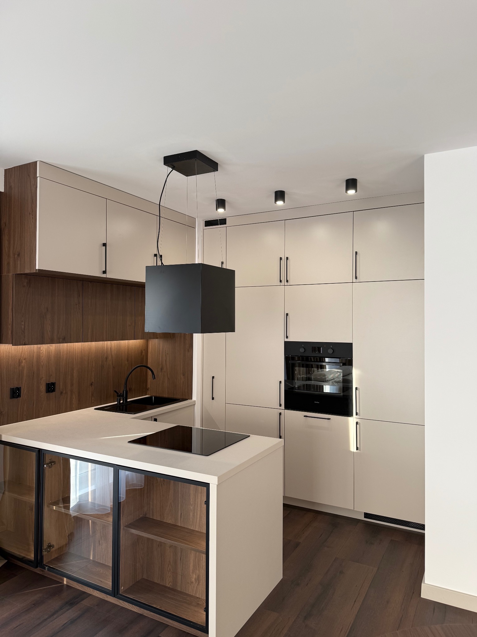 Nowoczesna kuchnia z wyspą, fronty w odcieniach beżu i drewna, czarne akcenty: bateria, okap, piekarnik. Oświetlenie punktowe podkreśla minimalistyczny design.