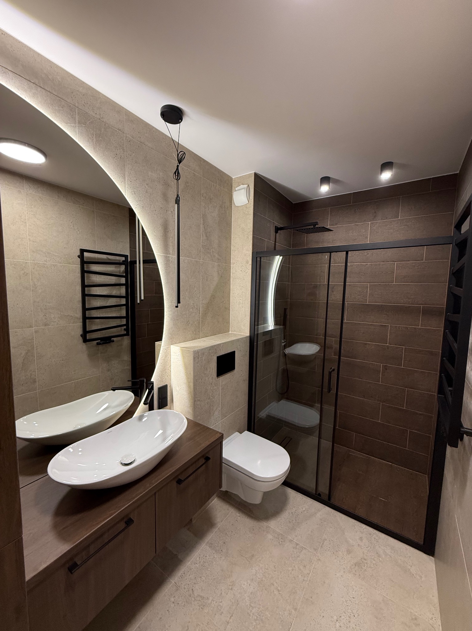 Nowoczesna łazienka z kabiną prysznicową typu walk-in, umywalką nablatową na drewnianej szafce i okrągłym lustrem z podświetleniem LED. Beżowo-brązowa kolorystyka.